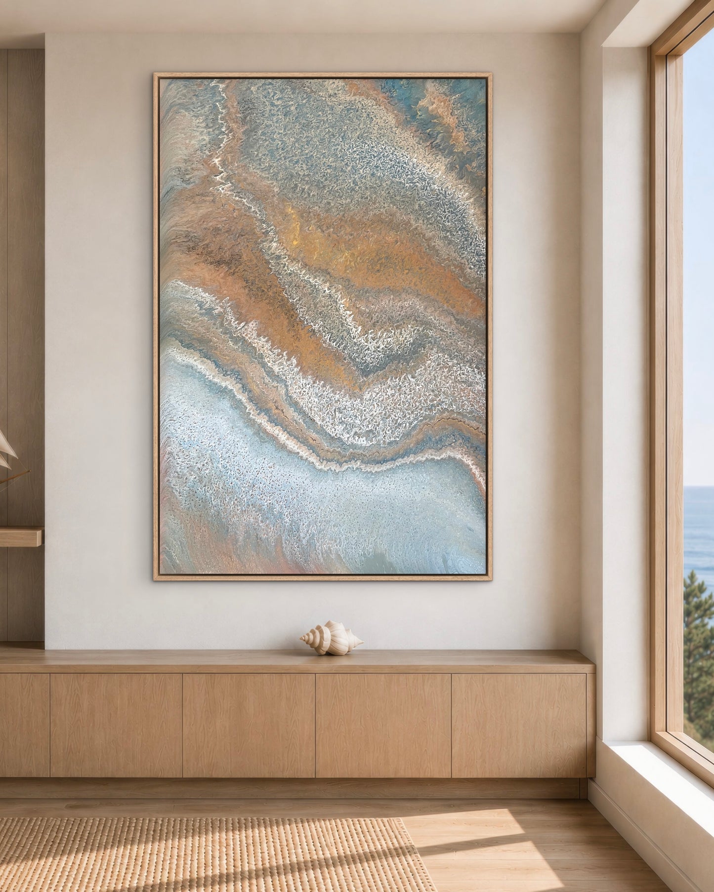 Weathered Tides blue and bronze coastal abstract painting displayed vertically above a light timber cabinet beside an ocean-view window
